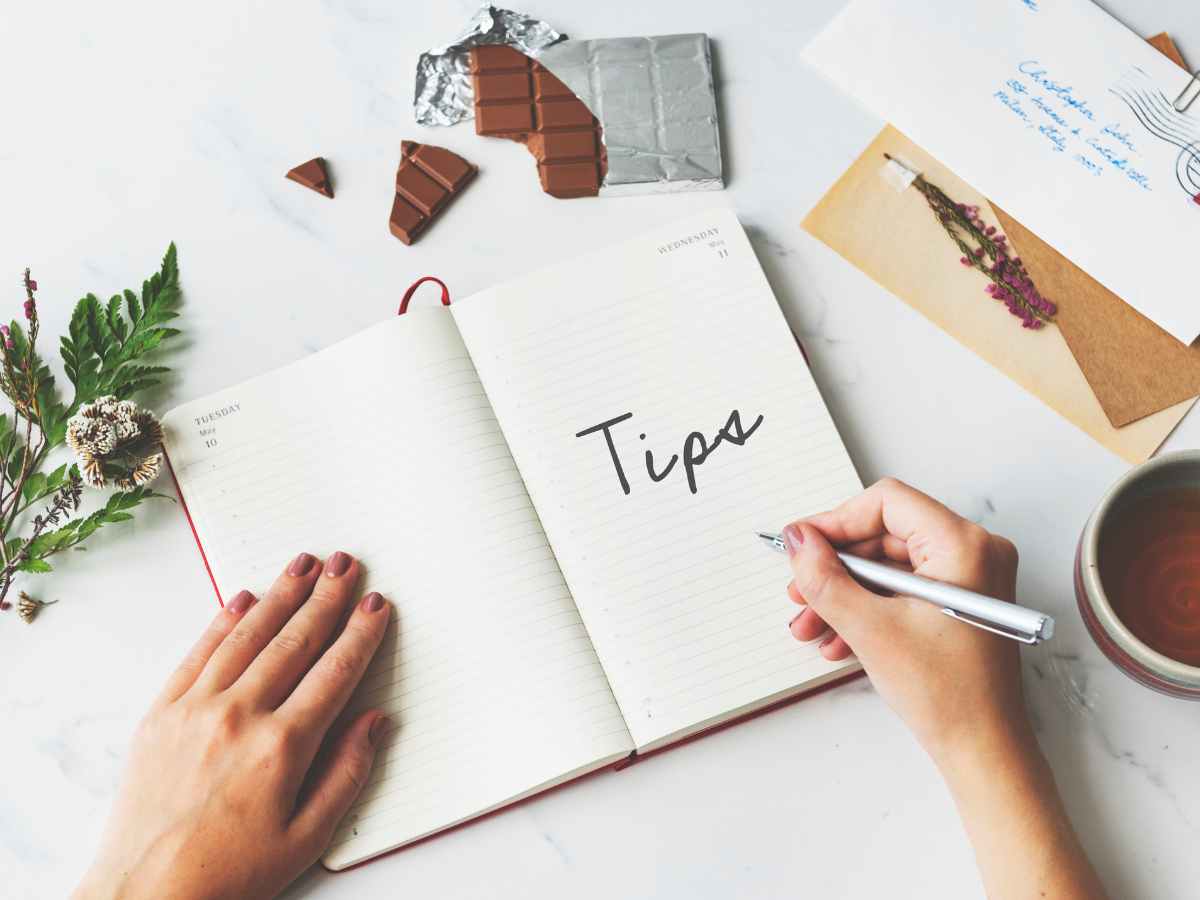 Pessoa escrevendo a palavra dicas em caderno aberto sobre mesa com chá, chocolate e cartas, simbolizando conselhos para projetos de mestrado.