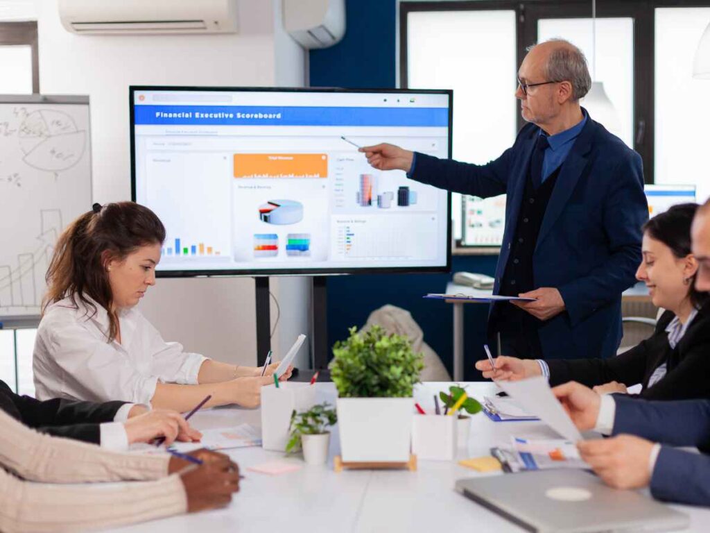 Professor apresentando dados financeiros em uma tela durante reunião com equipe.
